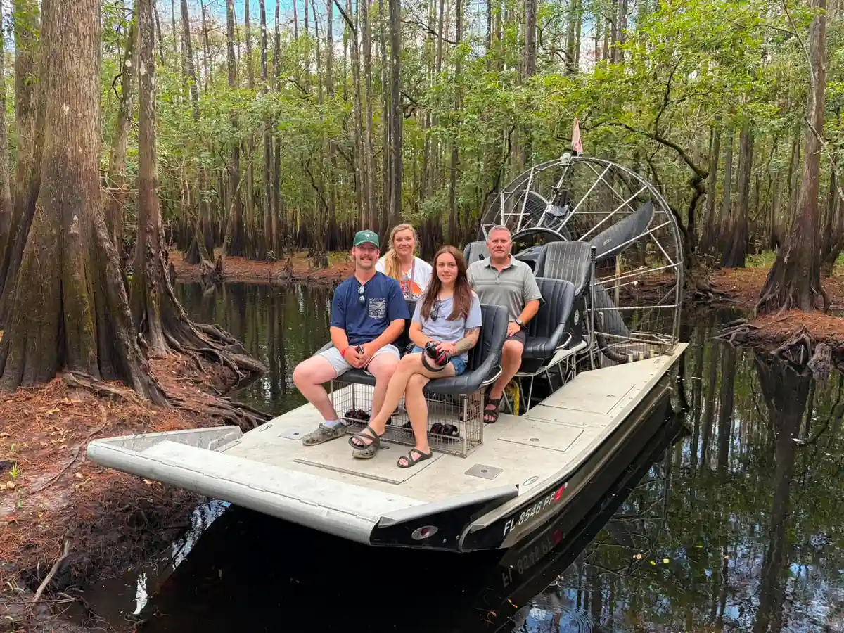 A family of four seated on an airboat deep in a shaded cypress slough, one holding a camera