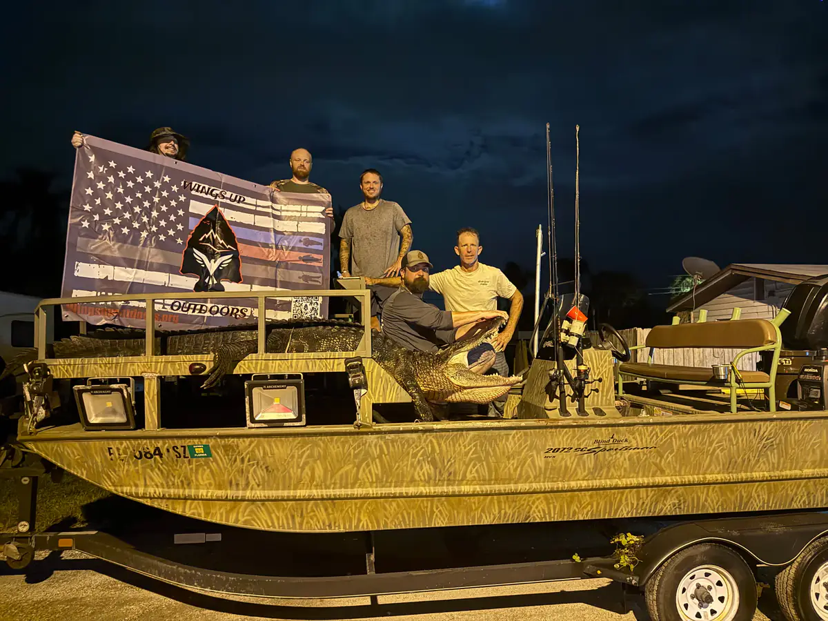 Five bow fishermen stand on a camouflage wrapped boat holding a flag at dusk before heading out