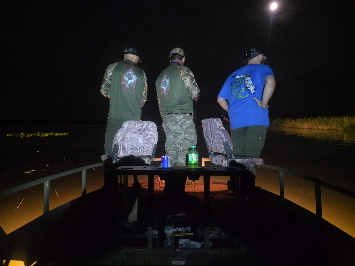 Three bow fishermen stand along the lit front deck scanning the lily pads for fish under a full moon