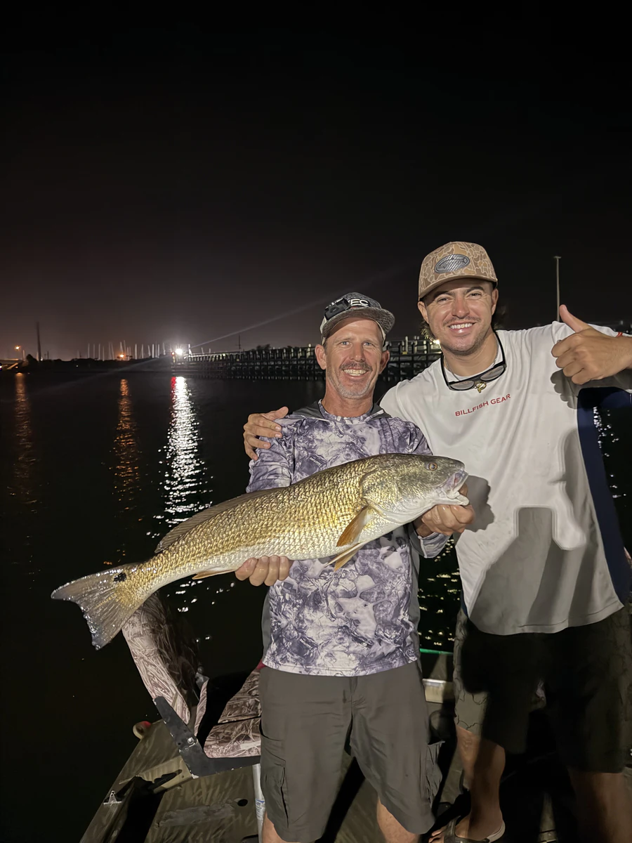 Two anglers holding up a large bronze redfish on the boat at night with causeway lights glowing behind them